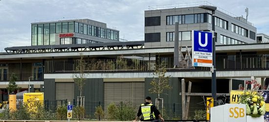 Budova s moderní architekturou a zelenými plochami, viditelná zastávka metra označená písmenem "U". V popředí se nachází pracovník v reflexní vestě.