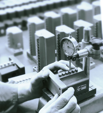 Ein Arbeiter überprüft mit einem Messgerät die Abmessungen eines Werkstücks in einer industriellen Umgebung. Im Hintergrund sind mehrere gestapelte, rechteckige Teile zu sehen.