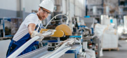 Ein Arbeiter in Schutzkleidung und Helm bedient eine Maschine in einer Industrieumgebung, während er ein langes Materialstück bearbeitet. Im Hintergrund sind weitere Maschinen und Materialien sichtbar.
