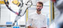 A man in a white shirt stands in a factory setting, observing a robotic arm. The robot is attached to a yellow structure and appears to be part of an industrial automation process.
