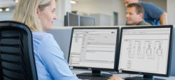 A woman is seated at a desk, working on a computer with two monitors. One monitor displays software interface data, while the other shows technical diagrams. In the background, a man is standing and observing her work. The setting appears to be an office environment.
