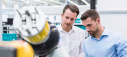 Two men are examining a robotic arm in a modern industrial setting, discussing its features and functionality. The background shows a well-lit workspace with various equipment.
