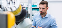 A man in a light blue shirt observes a robotic arm in a laboratory environment, focusing intently on the device.