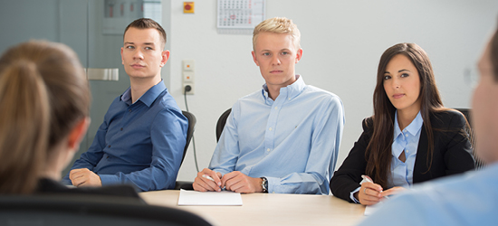 A group of four professionals is seated around a conference table, attentively listening to a speaker. Two men in light blue shirts and one woman in a black blazer are in the foreground, while another person is partially visible in the background. The setting appears to be a modern office environment.