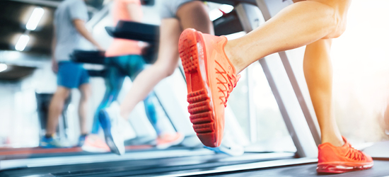 A close-up of a person's foot in bright red athletic shoes while running on a treadmill, with other gym-goers visible in the background. The setting is a bright indoor gym.