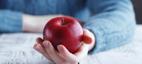 A hand holding a shiny red apple is placed on a textured surface, with a person's arm visible in the background, partially covered by a light blue sweater.