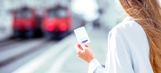 A person holding a white card stands in front of blurred red trains on a railway platform. The focus is on the card and the person's hand, while the trains are out of focus in the background.